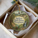 A round ornament with a scenic forest landscape and a trail sign, placed in a box with a white tissue paper lining.