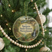 A round ornament hanging on a Christmas tree, featuring a scenic landscape with pine trees, a waterfall, and a trail marker.