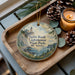 A wooden tray with a decorative ornament featuring a lighthouse and a scenic landscape, accompanied by pine cones and a lit candle.