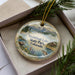 A decorative ornament in a box, featuring a scenic landscape with mountains, water, and pine trees.