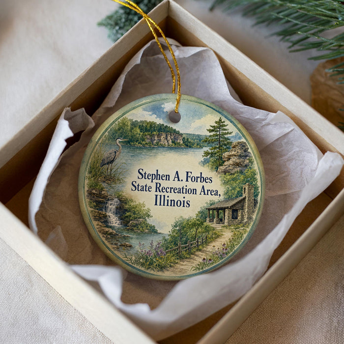 A decorative ornament in a white box, featuring a scenic landscape with a lake, mountains, and a bird.