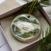 A decorative ornament with a scenic landscape featuring a lake, mountains, and a cabin, placed inside a box with pine branches.