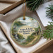 A round ornament with a scenic landscape featuring a waterfall, a bird, and a cabin, hanging from a string.