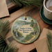 A round ornament with a scenic landscape depicting a waterfall, cabin, and forest, hanging on a wooden surface.