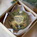 A decorative ornament in a box, featuring a scenic landscape with a waterfall, a cabin, and a deer.