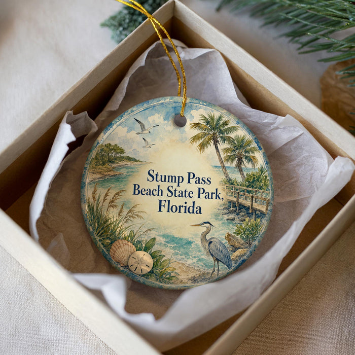 A round ornament with a beach scene and palm trees, placed in a box with a white tissue paper lining.