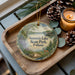 A decorative ornament with a scenic landscape of Summit Lake State Park, Indiana, featuring a deer, pine trees, and a lake. The ornament is placed on a wooden tray, with pine cones and a lit candle nearby.