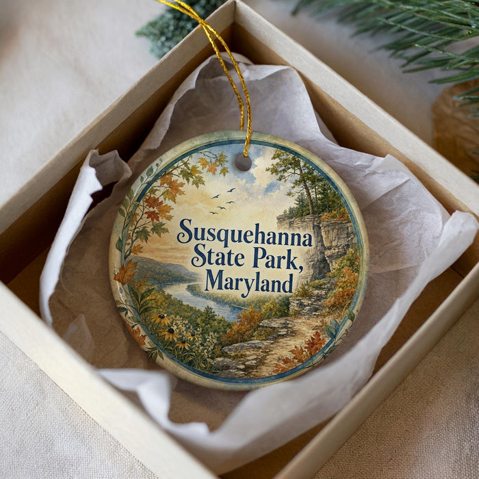 A decorative ornament in a box, featuring a scenic landscape with a river, trees, and a path leading to Susquehanna State Park, Maryland.