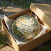 A decorative ornament in a box, featuring a scenic landscape with trees, a river, and a castle, with the text "Susquehanna State Park, Maryland" printed on it.