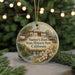 A round ornament with a painting of a historical building and a river, surrounded by greenery, hangs in front of a blurred background with twinkling lights.
