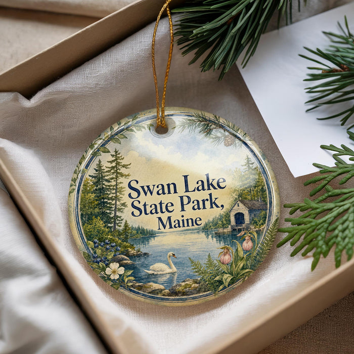A decorative ornament with a scenic lake and forest landscape, featuring a swan and a cabin, is displayed in a box with pine branches.