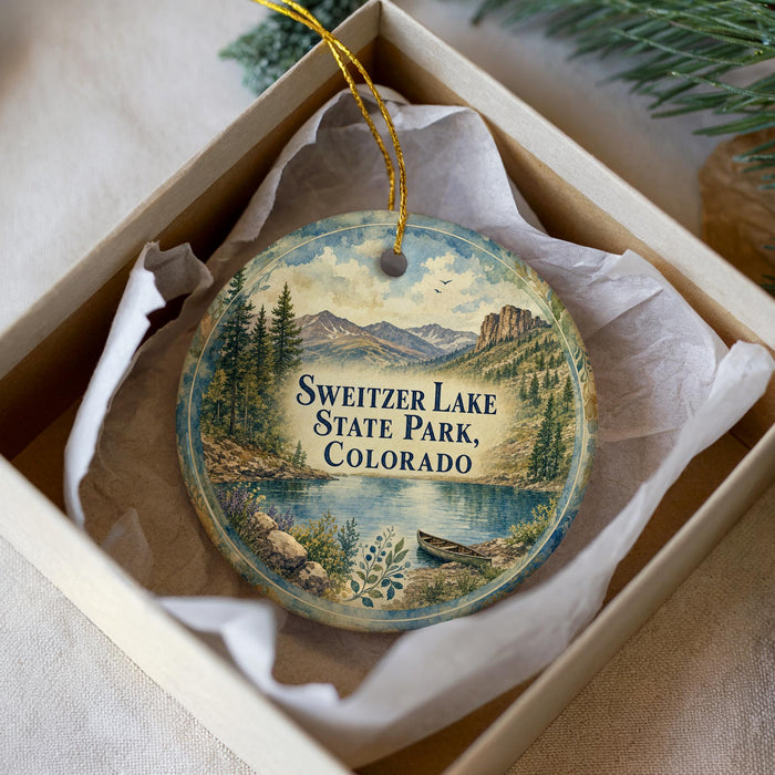 A decorative ornament in a box, featuring a scenic landscape with mountains, trees, and a lake.