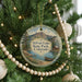 A round ornament hanging on a Christmas tree, featuring a scenic landscape with mountains, a lake, and a cabin.