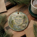 A decorative ornament featuring a scenic landscape with mountains, pine trees, and a river, hanging on a wooden surface.