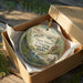 A decorative ornament in a wooden box, featuring a scenic landscape with a river, mountains, and a covered wagon, along with the text "Three Island Crossing State Park, Idaho".