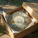 An oval ornament in a brown box, featuring a scenic landscape with mountains, trees, and a waterfall.