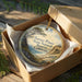 A round ornament in a box, featuring a scenic landscape with a pine tree and mountains, with the text "Torrey Pines State Natural Reserve, California" printed on it.