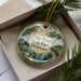 A decorative ornament in a box, featuring a scenic image of a pond with a bird and a person in a boat, along with the text " Trap Pond State Park, Delaware".