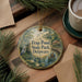 A round ornament with a painting of a bird and a pond, surrounded by pine branches, sits on a wooden surface.
