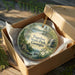 A decorative ornament in a wooden box, featuring a scenic landscape with a waterfall and pine trees, with the text "Tugaloo State Park, Georgia" displayed on the ornament.