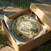 A round ornament in a box, featuring an illustration of Upper Sioux Agency, State Park, and Minnesota, with a tiki hut and other natural elements.