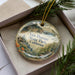 A decorative ornament in a box, featuring a scenic landscape with a house and pine trees, along with the text "Van Damme State Park, California".