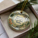 A decorative ornament in a box, featuring a scenic landscape with mountains, a waterfall, and a monument, with the text "Veterans Memorial State Park, Idaho" prominently displayed.