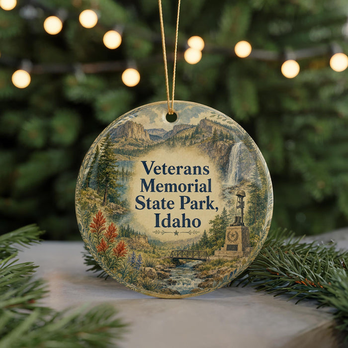 A round ornament with a scenic landscape and text that reads "Veterans Memorial State Park, Idaho" hangs in front of a blurred background of Christmas lights.
