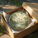 A decorative ornament in a wooden box, featuring a scenic landscape with a river, trees, and a boat.