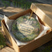 A circular ornament with a scenic image of Wadsworth Falls State Park in Connecticut, placed in a wooden box with a gold string.