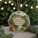 A circular ornament with a tropical landscape design, featuring a river, palm trees, and a house, is displayed in front of a blurred background with Christmas lights.