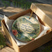 A circular ornament with a tropical landscape design, placed in a cardboard box with a gold string.