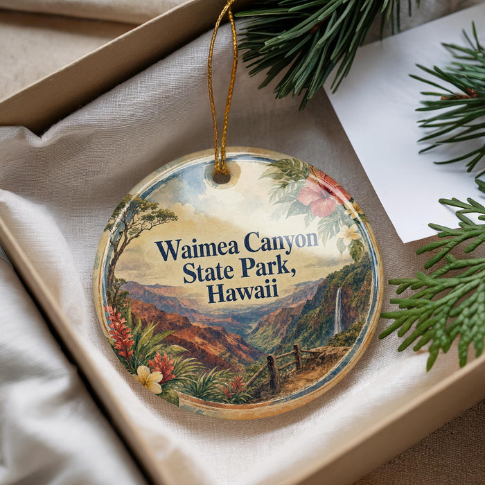 A decorative ornament with a scenic image of Waimea Canyon State Park, Hawaii, is displayed in a box with pine branches and a white card.