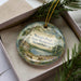 A decorative ornament in a box, featuring a scenic landscape with a pond, trees, and a cabin.