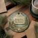 A round ornament with a scenic landscape of Walden Pond, featuring a cabin and pine trees, is displayed on a wooden surface alongside a cup of coffee and some papers.