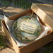A round ornament in a box, featuring a scenic landscape with a pond, trees, and a cabin, is displayed on a wooden surface.