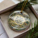 A decorative ornament in a box, featuring a scenic landscape with mountains, trees, and a deer.
