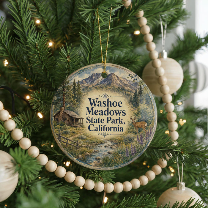 A round ornament hanging on a Christmas tree, featuring a scenic landscape with mountains, a cabin, and a deer.