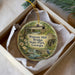 A round ornament with a painting of a bridge and flowers, placed in a box with a white tissue paper lining.