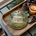 A decorative ornament featuring a scenic landscape of Wauboardsie State Park, Iowa, with a bird perched on a tree branch. The ornament is displayed on a wooden tray, accompanied by pine cones and a lit candle, creating a cozy and festive atmosphere.