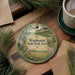 A decorative ornament featuring a scenic landscape of Waubonsie State Park, Iowa, with a bird perched on a tree branch. The ornament is placed on a wooden surface, surrounded by pine branches and a cup of coffee.