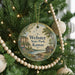 A round ornament hanging on a Christmas tree, featuring a scenic landscape with a river, trees, and a cabin.