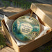 A decorative ornament in a box, featuring a mermaid and a tropical landscape, with the text "Weeki Wachee Springs State Park, Florida" printed on it.