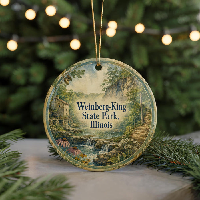 A circular ornament with a scenic landscape of a waterfall and a house, surrounded by greenery, is displayed in front of a blurred background with twinkling lights.