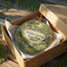 A round ornament in a box, featuring a scenic landscape with a house and trees, is displayed on a wooden surface.