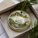 A decorative ornament in a box, featuring a scenic landscape with a waterfall and pine trees.