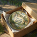 A decorative ornament in a brown box, featuring a scenic landscape with a waterfall and pine trees, with the text "Whewater State Park, Minnesota" printed on it.