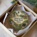 A decorative ornament in a box, featuring a scenic landscape with a waterfall, trees, and a cabin, with the text "Wildcat Den State Park, Iowa" printed on it.