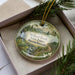 A decorative ornament in a box, featuring a scenic landscape with a waterfall, pine trees, and a path leading through the forest.