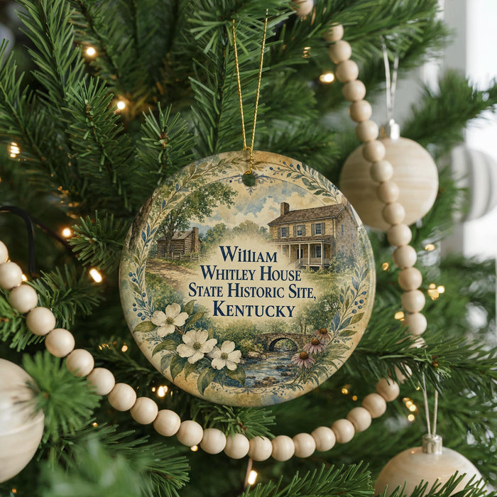 A round ornament hanging on a Christmas tree, featuring a painting of a house surrounded by flowers and greenery.